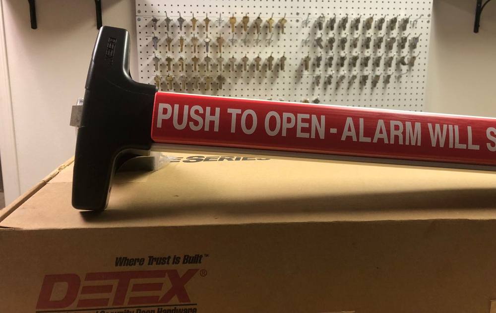 A Detex panic bar marked "Push to open, alarm will sound", in front of a board of key blanks
